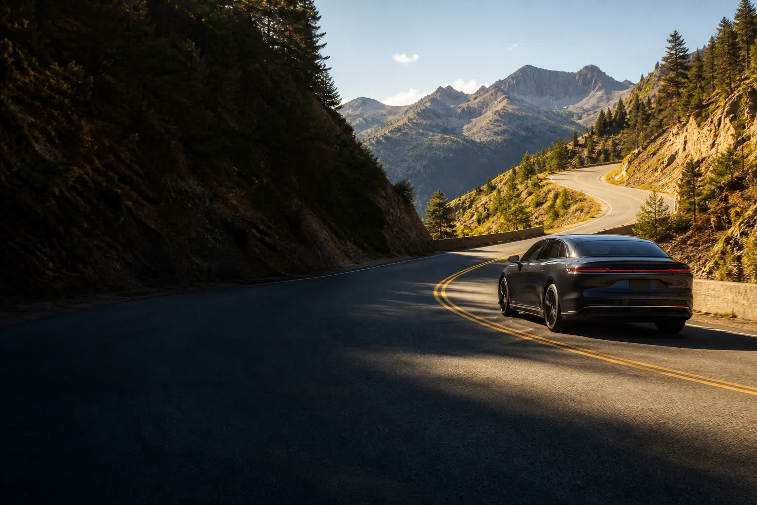 A sunlit winding mountain road with a dark electric car on the right.